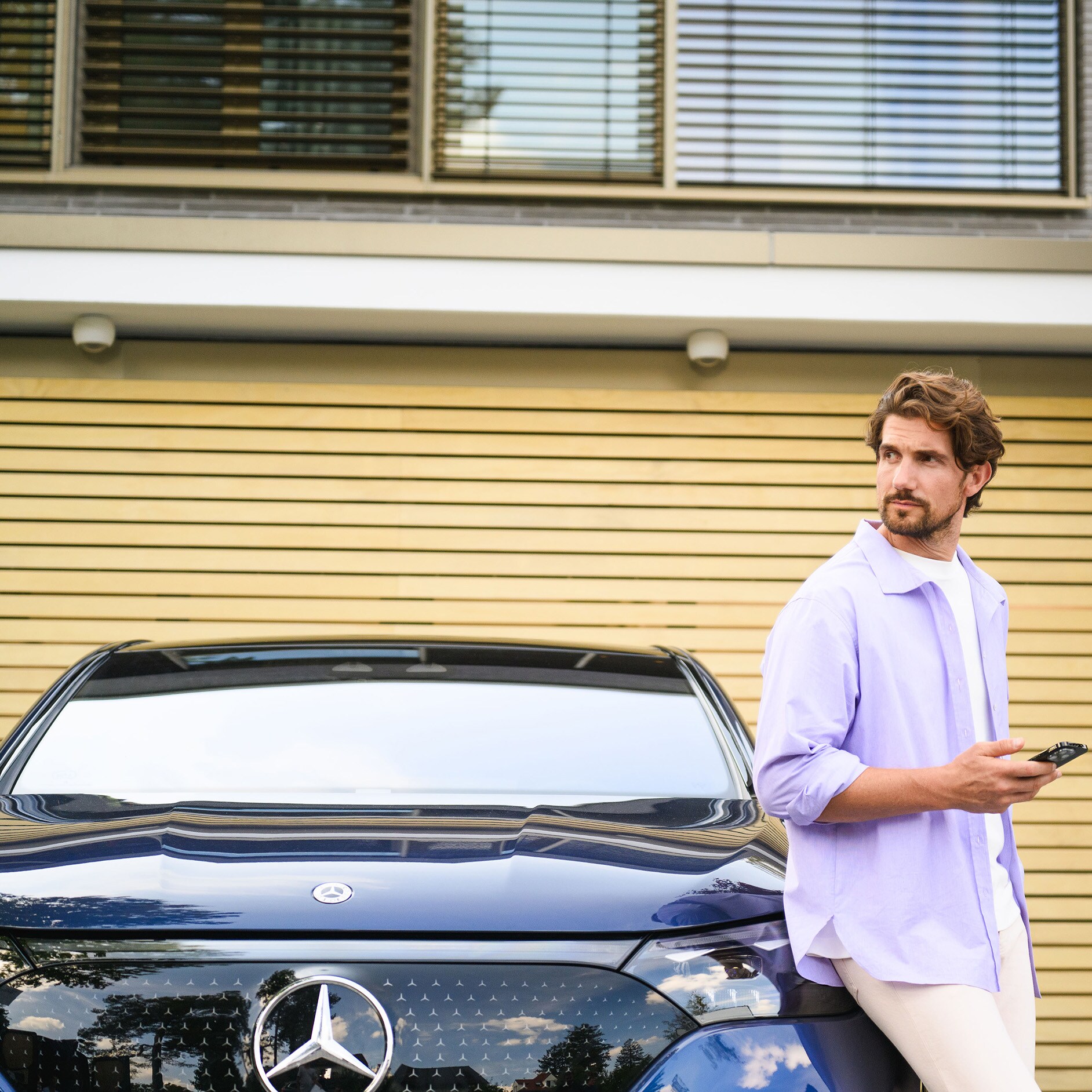 Financieringsaanbod voor uw tweedehands Mercedes-Benz Een persoon leunend tegen een Mercedes-Benz voertuig, kijkend in de verte terwijl hij een mobiel apparaat vasthoudt.