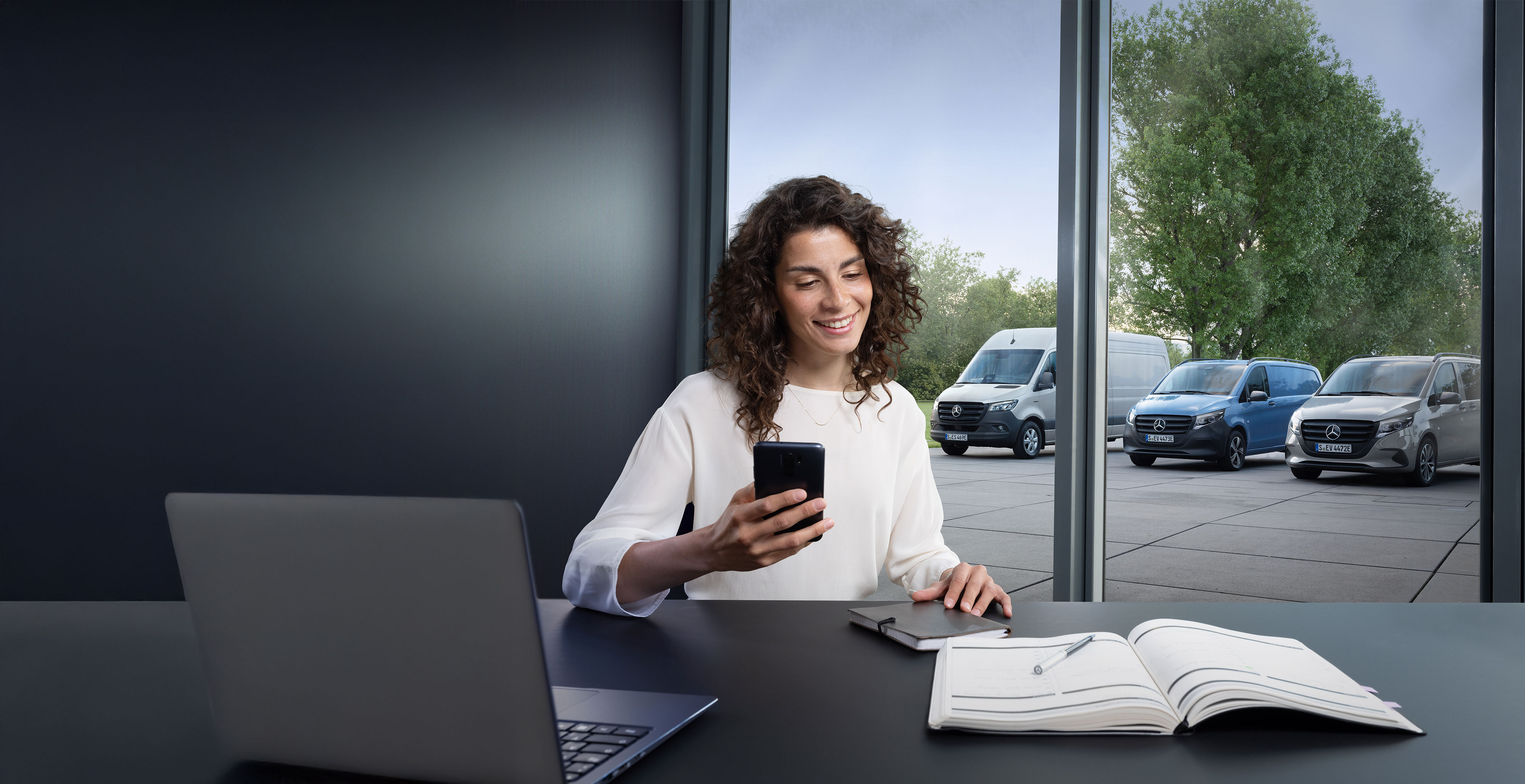 Mercedes-Benz | Confort | Prise de rendez-vous en ligne Femme réservant un service par smartphone avec une gamme d’utilitaires léger Mercedes-Benz en arrière-plan.